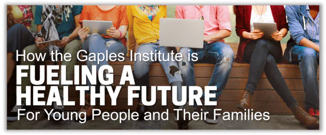 A diverse group of young people sitting on a bench. One is looking at a laptop, another is looking at a tablet.